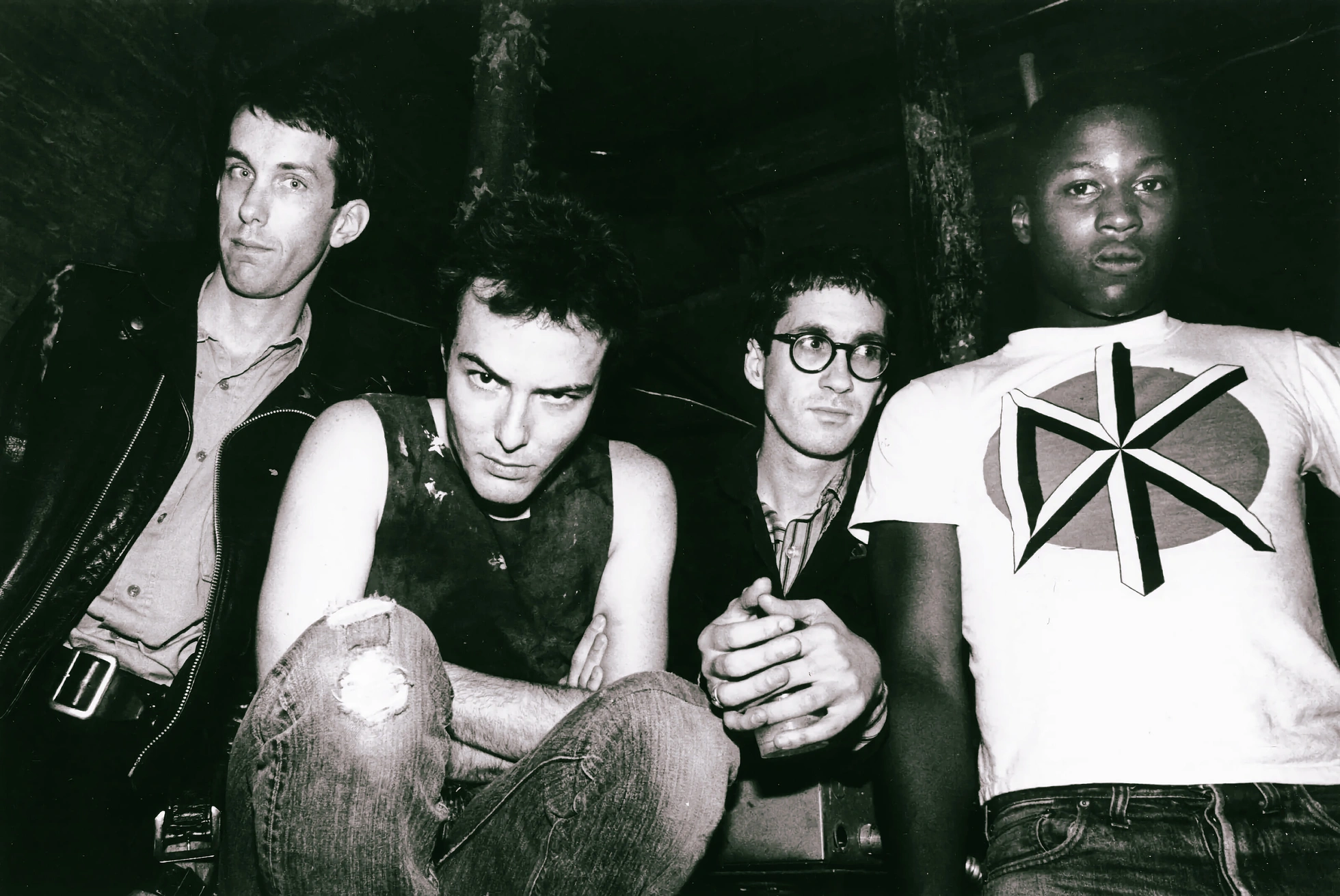 A black-and-white photograph of the Dead Kennedys, the four band members posed together in a dim room, one wearing a t-shirt printed with the band's DK logo.