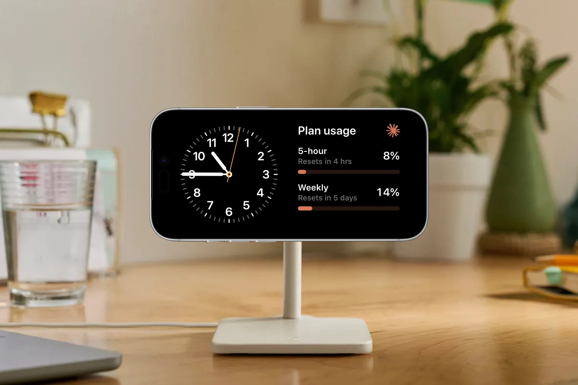 An iPhone in landscape on a MagSafe stand on a wooden desk, displaying iOS StandBy mode with an analog clock on the left and the claudeusage Plan usage widget on the right tracking 5-hour and weekly limits. A glass of water and a plant sit in the background, with the corner of a MacBook in the foreground.