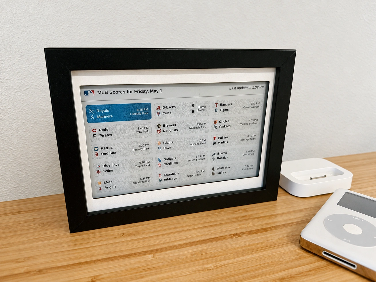 The e-ink scoreboard in a frame on a desk, showing a 3x5 grid of live MLB scores in dithered six-color rendering.