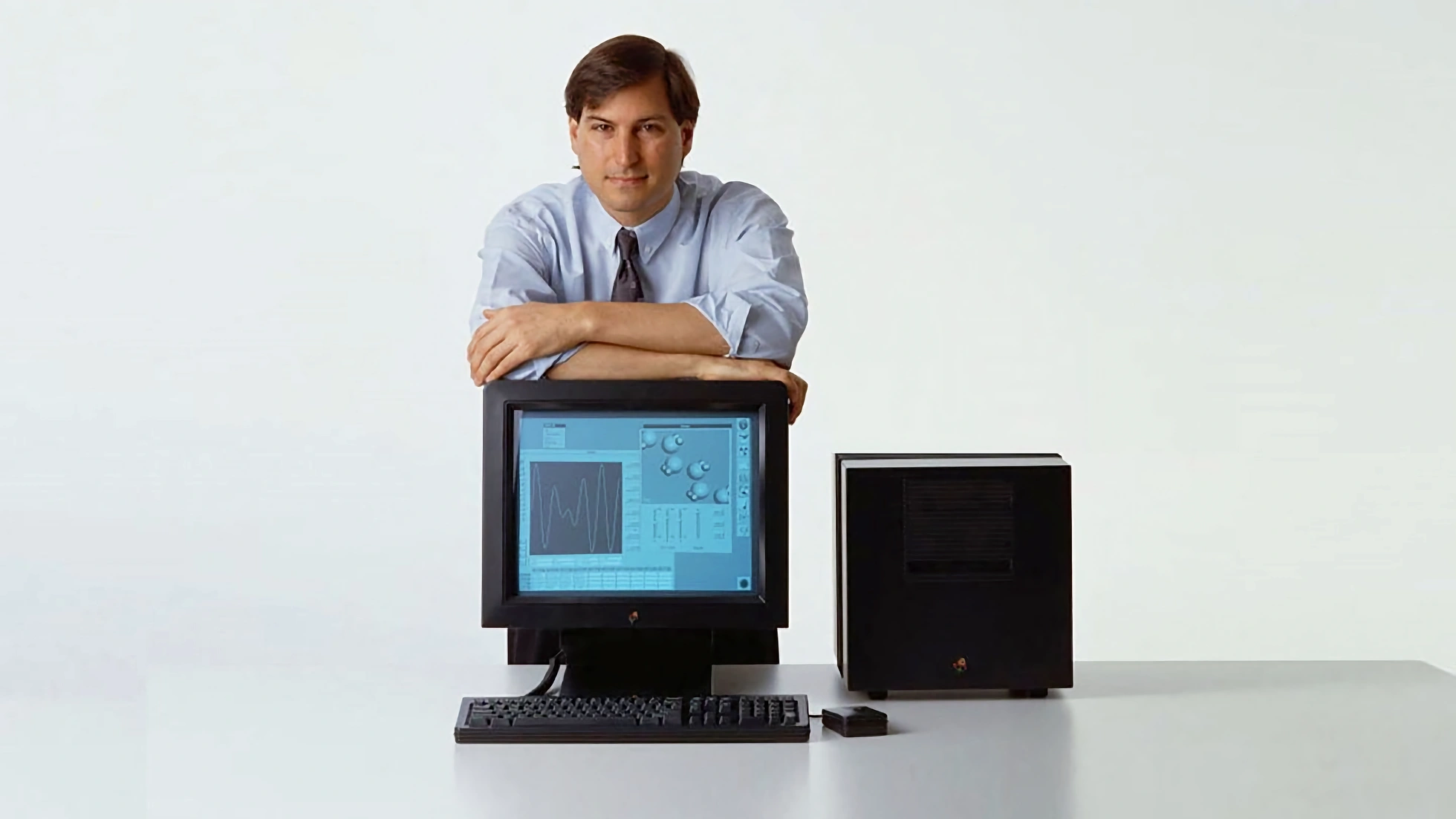 Steve Jobs leaning on a NeXT MegaPixel monitor with a NeXTcube to its right, shot in studio lighting against a near-white background. Matte black magnesium housings, discrete keyboard and mouse.