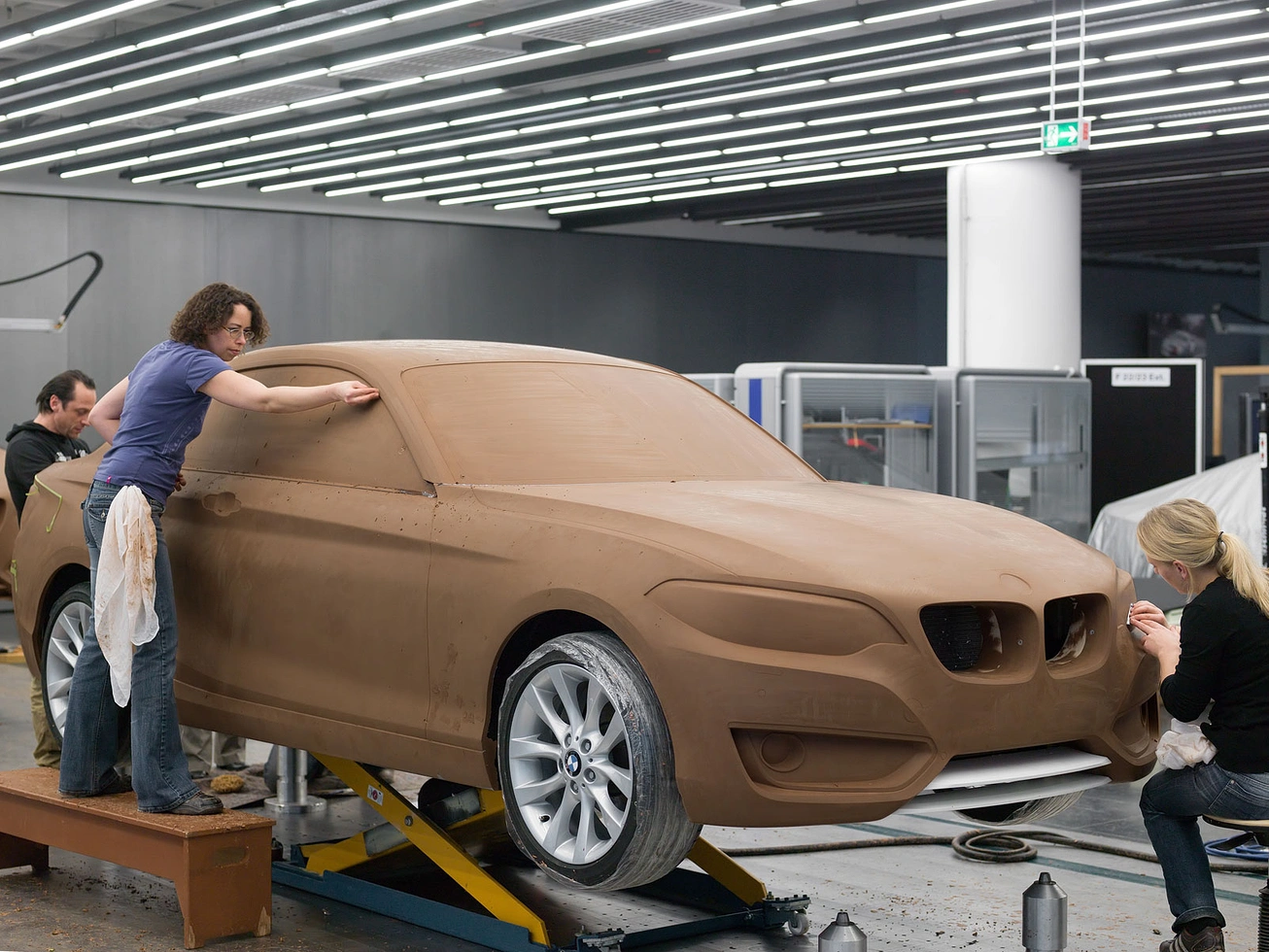A full-size BMW 2 Series coupe sculpted in raw, unpainted clay, mounted on a platform with real production wheels attached. Three sculptors work on it: one leans over the roof smoothing the surface with a cloth, one on the far side refines the A-pillar, and a third crouches on the passenger side detailing the headlight with a handheld tool. Bright linear ceiling lights fill the studio.