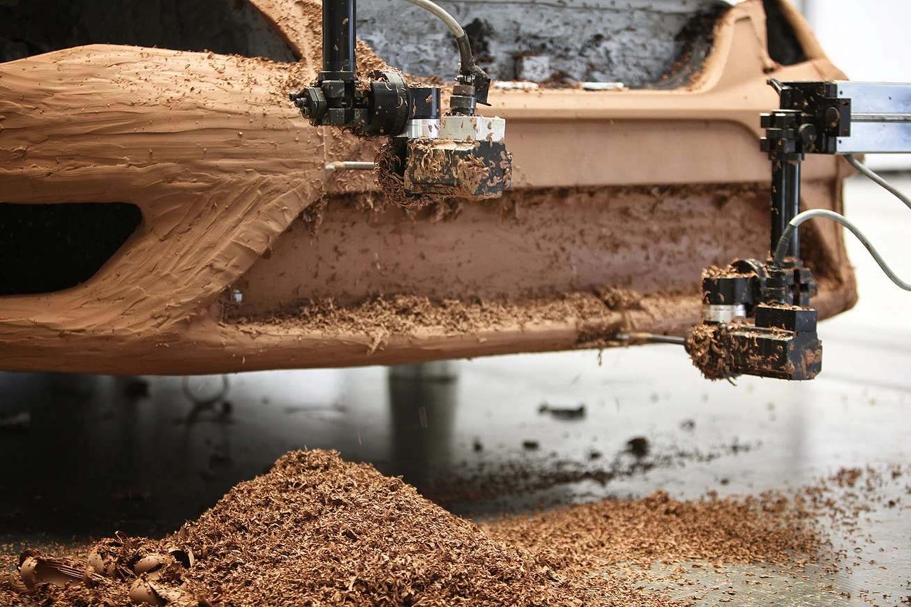 Close-up of two black robotic CNC arms milling a full-size clay model of a Seat Leon. Thick curls of brown clay fly off the cutter heads and pile up on the studio floor below. The rough shape of a front fender and bumper is visible, covered in striated tool marks.