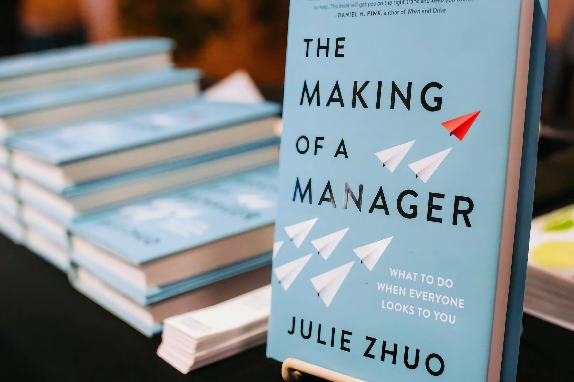 Julie Zhuo's book The Making of a Manager propped upright on a book-signing table, with a stack of copies behind it in soft focus. The cover shows a pale blue background, the title in bold black serif, and a cluster of paper airplanes — one red, the rest white — flying toward the top right.
