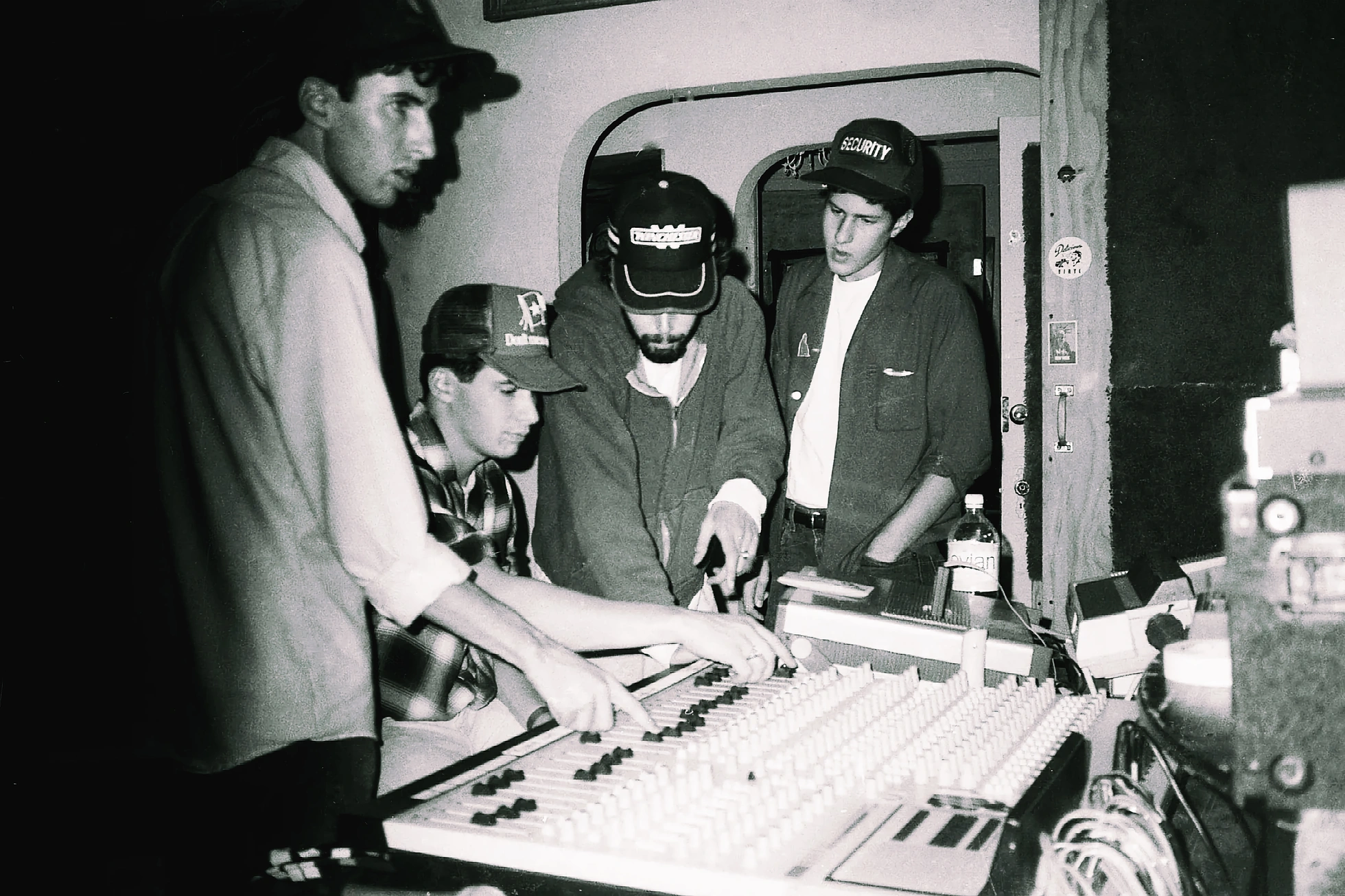 Black-and-white studio photograph of four young men crowded around a mixing console. Two of them have hands on the faders, the others lean in watching. Three wear baseball caps, one with the word SECURITY across the front. Outboard gear and cabling are visible behind them.