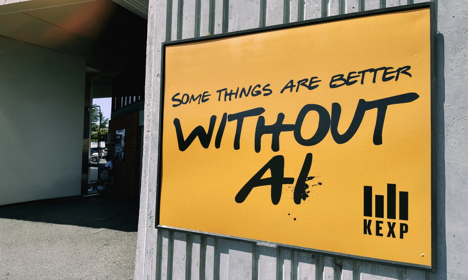 A yellow KEXP outdoor billboard mounted on a corrugated wall, reading SOME THINGS ARE BETTER WITHOUT AI in handwritten black brush ink, with the KEXP logo in the bottom right corner.