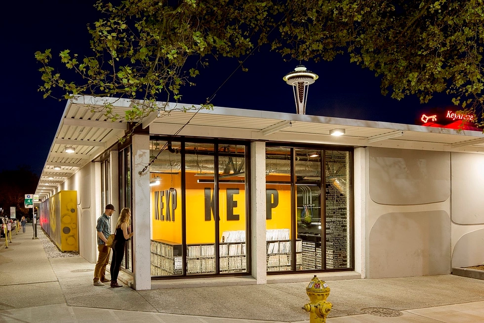 KEXP's Seattle Center studios illuminated at night, with people looking through the record-filled windows and the Space Needle rising above the roofline.