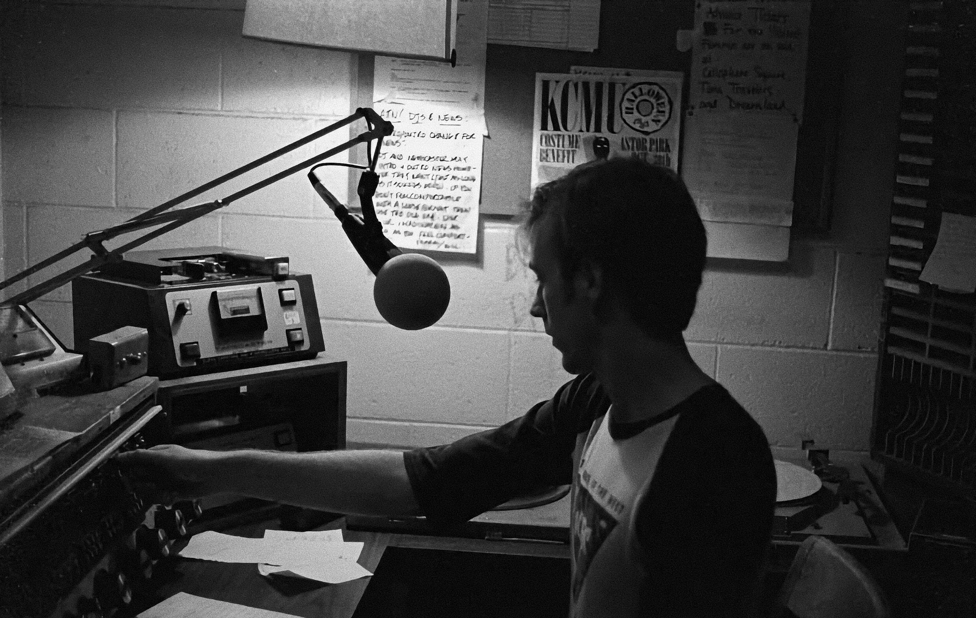 Black-and-white photograph of a young DJ at the KCMU radio studio mid-1980s, reaching for a knob on the mixing board with a boom-mounted microphone in front of him. A KCMU Halloween benefit poster is taped to the cinderblock wall behind him next to handwritten DJ notes; a reel-to-reel deck sits on a shelf nearby.