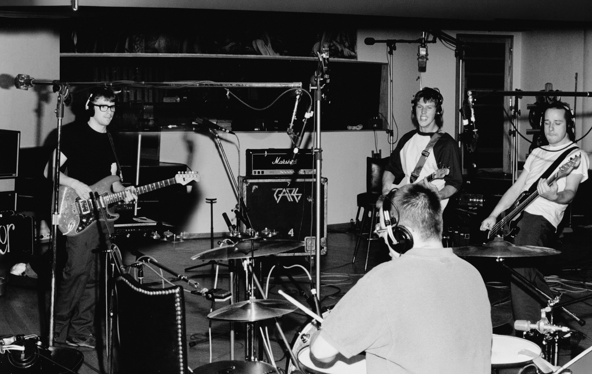 Black-and-white photograph of Weezer recording in a studio. Rivers Cuomo on the left in a t-shirt and glasses plays guitar at a microphone. Brian Bell stands in the middle with a guitar; Matt Sharp is on the right holding a bass. Pat Wilson sits at the drum kit in the foreground, back to the camera, wearing headphones. Marshall amps and recording gear fill the room.